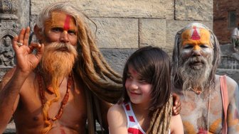 Ein Mädchen mit langen Haaren sitzt zwischen zwei heiligen Männern, die farbenfrohe Gesichtsbemalung tragen und freundlich lächeln.