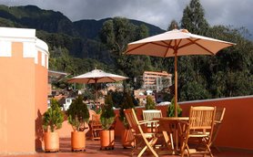 Eine sonnige Terrasse mit Holzmöbeln und Sonnenschirmen, umgeben von grünen Pflanzen und einer beeindruckenden Berglandschaft.