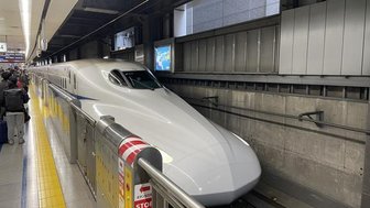 Ein moderner Shinkansen-Zug steht an einem Bahnhof, bereit zur Abfahrt, während Passagiere in der Nähe warten.