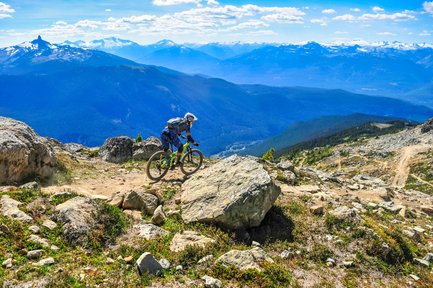 Ein Mountainbiker fährt über einen steinigen Pfad mit beeindruckender Berglandschaft im Hintergrund.
