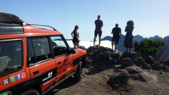 Ein orangefarbener Jeep steht neben einer Gruppe von vier Personen, die auf einer Klippe die Aussicht genießen.