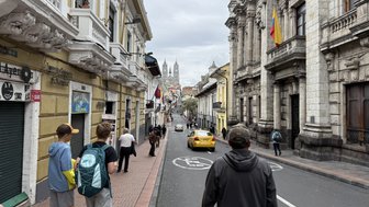 Zwei Jugendliche gehen die Straße entlang, während ein gelbes Taxi vorbeifährt und historische Gebäude im Hintergrund sichtbar sind.