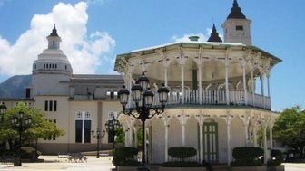 Ein eleganter Pavillon mit Säulen steht im Zentrum eines sonnigen Platzes, umgeben von Bäumen und historischen Gebäuden.