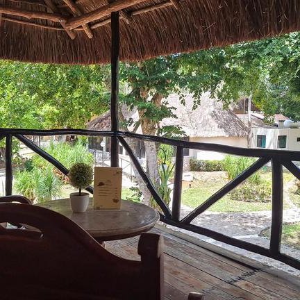 Ein gemütlicher Balkon mit einem Holzstuhl und Tisch, umgeben von üppigem Grün und traditionellen Hütten im Hintergrund.