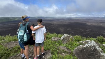 Eine Mutter steht mit zwei Kindern an einem Aussichtspunkt und blickt auf eine weite, vulkanische Landschaft.