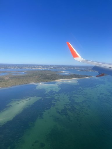 Das Bild zeigt den Blick aus einem Flugzeugfenster auf eine malerische Küstenlinie und glitzerndes Wasser.