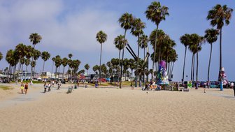 Ein belebter Strand mit Palmen, wo Menschen spazieren gehen und sich entspannen, während Graffiti-Kunst im Hintergrund sichtbar ist.