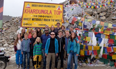 Eine Gruppe von Reisenden posiert fröhlich vor dem Khardungla-Pass-Schild, umgeben von bunten Gebetsfahnen.