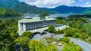 Ein modernes Hotel mit grünem Dach, umgeben von üppiger Natur und Blick auf einen ruhigen See und die Berge im Hintergrund.