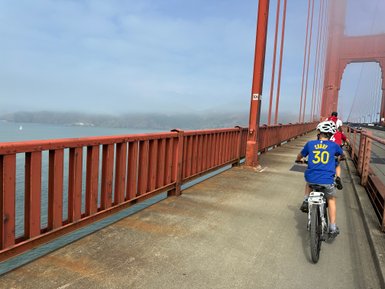 Ein Junge in einem blauen T-Shirt fährt mit dem Fahrrad über die Golden Gate Bridge, umgeben von Nebel und Wasser.