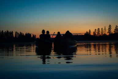 Vier Personen sitzen in Kanus auf einem ruhigen See, während die Sonne hinter den Bäumen untergeht und den Himmel färbt.