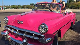 Zwei Frauen stehen lächelnd neben einem leuchtend pinken Oldtimer-Cabrio unter einem strahlend blauen Himmel.