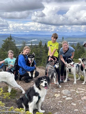Eine Gruppe von Menschen posiert fröhlich mit mehreren Huskys auf einem Berg, umgeben von einer malerischen Landschaft.