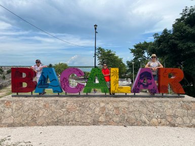Drei Personen posieren fröhlich vor einem bunten 'Bacalar'-Schild, umgeben von einer malerischen Landschaft und blauem Himmel.