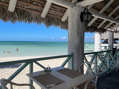Ein Blick auf einen Strand mit klarem Wasser, wo Kinder im seichten Wasser spielen, während ein Restaurant im Hintergrund sichtbar ist.