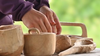 Eine Hand formt sorgfältig ein Holzobjekt, während mehrere geschnitzte Holzgegenstände auf einem Tisch liegen.
