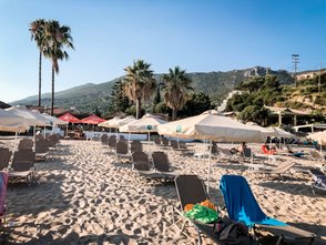 Ein sonniger Strand mit Liegen und Sonnenschirmen, umgeben von Palmen und sanften Hügeln im Hintergrund.
