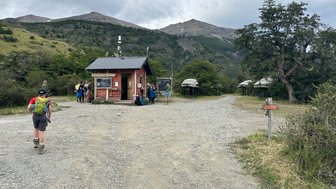 Ein Wanderer mit Rucksack geht auf einem Schotterweg zu einer kleinen roten Hütte in der Natur Patagoniens.