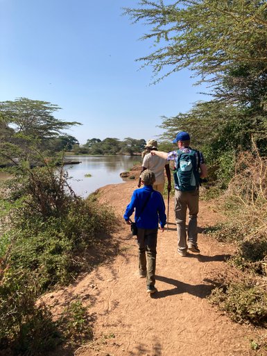 Eine Gruppe von drei Personen wandert entlang eines schmalen Pfades am Ufer eines ruhigen Gewässers unter klarem Himmel.