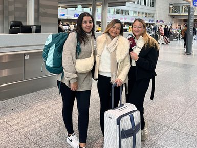 Drei Frauen stehen am Flughafen Frankfurt, lächelnd und bereit für ihre Reise, mit Gepäck und einem fröhlichen Ausdruck.