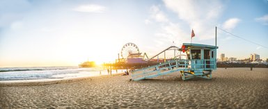 Ein Rettungsturm steht am Strand von Santa Monica, während die Sonne hinter dem Pier untergeht und die Wellen sanft an den Strand rollen.