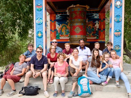 Eine Reisegruppe sitzt lächelnd vor einem bunten tibetischen Tempel mit einem großen Gebetsrad im Hintergrund.