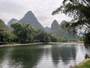 Ein ruhiger Fluss spiegelt die majestätischen, bewaldeten Karstberge unter einem bewölkten Himmel wider.