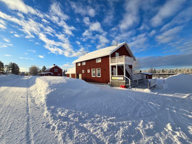 Ein rotes Holzhaus steht in einer schneebedeckten Landschaft unter einem strahlend blauen Himmel mit einigen Wolken.
