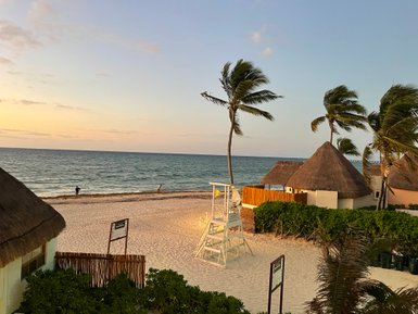 Ein ruhiger Strand mit sanften Wellen, umgeben von Palmen und traditionellen Strohdächern, während die Sonne untergeht.