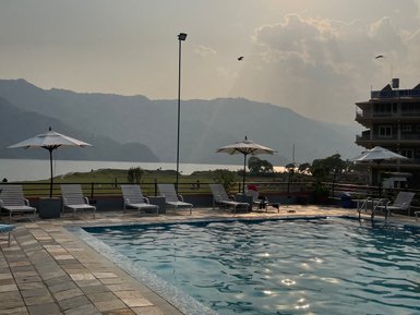 Ein Pool mit Liegen und Sonnenschirmen, umgeben von Bergen und einem ruhigen See, während die Sonne am Himmel scheint.