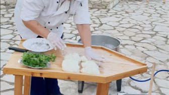 Eine Person in Kochkleidung bereitet frischen Halloumi-Käse auf einem Holztisch im Freien vor.