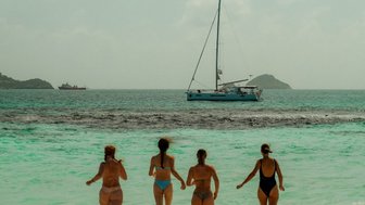 Vier Frauen in Bikinis laufen fröhlich ins Wasser, während ein Segelboot im Hintergrund sanft auf den Wellen schaukelt.