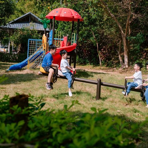 Vier Kinder spielen fröhlich auf einer Wippe im Freien, umgeben von grünem Gras und einem Spielplatz im Hintergrund.