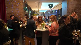 In einem eleganten Raum stehen Gäste an Stehtischen, während sie Getränke genießen und sich unterhalten.