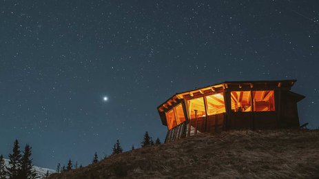 Eine beleuchtete Hütte steht auf einem Hügel, umgeben von dunklem Himmel und funkelnden Sternen.