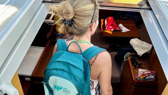 Eine Frau mit einem blauen Rucksack steigt eine Treppe auf einem Boot hinauf, während Sonnenlicht durch das Fenster strömt.