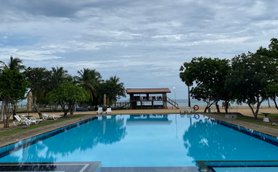 Ein weitläufiger Pool spiegelt den bewölkten Himmel wider, umgeben von Palmen und einer einladenden Liegewiese.