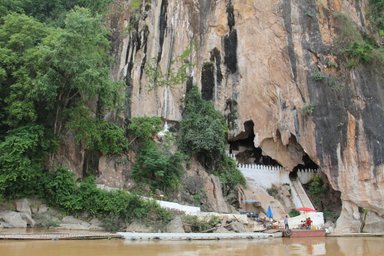 Eine beeindruckende Felswand mit üppigem Grün, die eine Höhle und eine Treppe zu einem Tempel zeigt.
