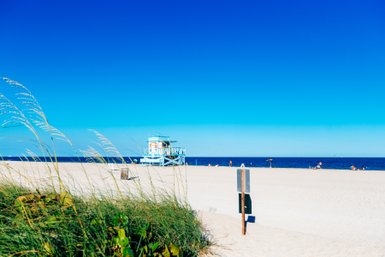 Ein sonniger Strand mit feinem Sand, einem blauen Rettungsturm und Menschen, die sich am Wasser entspannen.