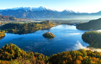 Ein malerischer Blick auf den Bleder See, umgeben von buntem Herbstlaub und majestätischen Bergen im Hintergrund.