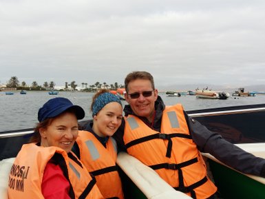Drei Personen in orangefarbenen Schwimmwesten lächeln auf einem Boot, umgeben von Wasser und Booten im Hintergrund.