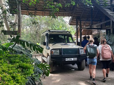Zwei Personen mit Rucksäcken gehen in Richtung eines überdachten Eingangs, während ein Geländewagen parkt.