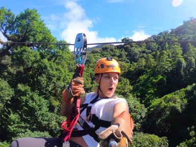 Eine Person mit einem orangefarbenen Helm schwingt fröhlich an einer Zipline über üppige grüne Bäume und strahlend blauem Himmel.