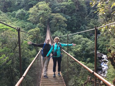 Zwei Wanderer stehen auf einer Hängebrücke, umgeben von üppigem Grün und einem Wasserfall im Hintergrund.