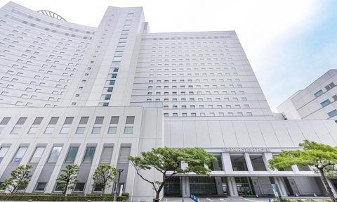 Ein modernes Hotelgebäude mit einer hellen Fassade und großen Fenstern, umgeben von gepflegten Bäumen und einem Parkplatz.