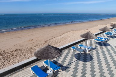 Ein ruhiger Strand mit sanften Wellen, blauen Liegen und Sonnenschirmen, die Schatten auf den gepflasterten Boden werfen.