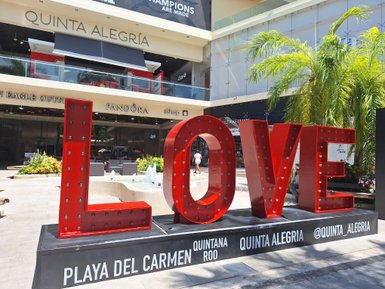Ein großes, leuchtendes 'LOVE'-Schild steht vor einem Einkaufszentrum in Playa del Carmen, umgeben von Palmen und Blumen.