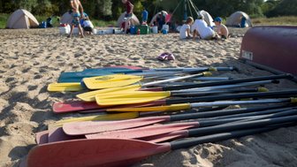 Bunte Paddel liegen im Sand, während Kinder und Erwachsene in der Nähe Zelte aufbauen und sich auf das Campen vorbereiten.