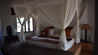 Ein gemütliches Schlafzimmer mit einem großen Bett, umgeben von einem Moskitonetz und dekorativen Kissen in warmen Farben.