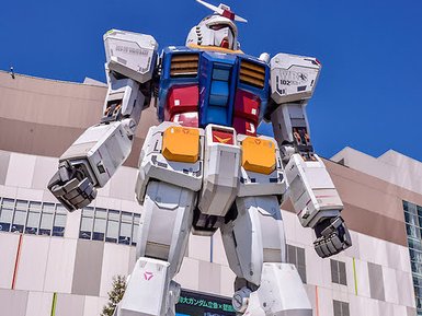Ein riesiger Gundam-Roboter steht majestätisch vor einem modernen Gebäude in Tokyo, umgeben von klarem blauen Himmel.